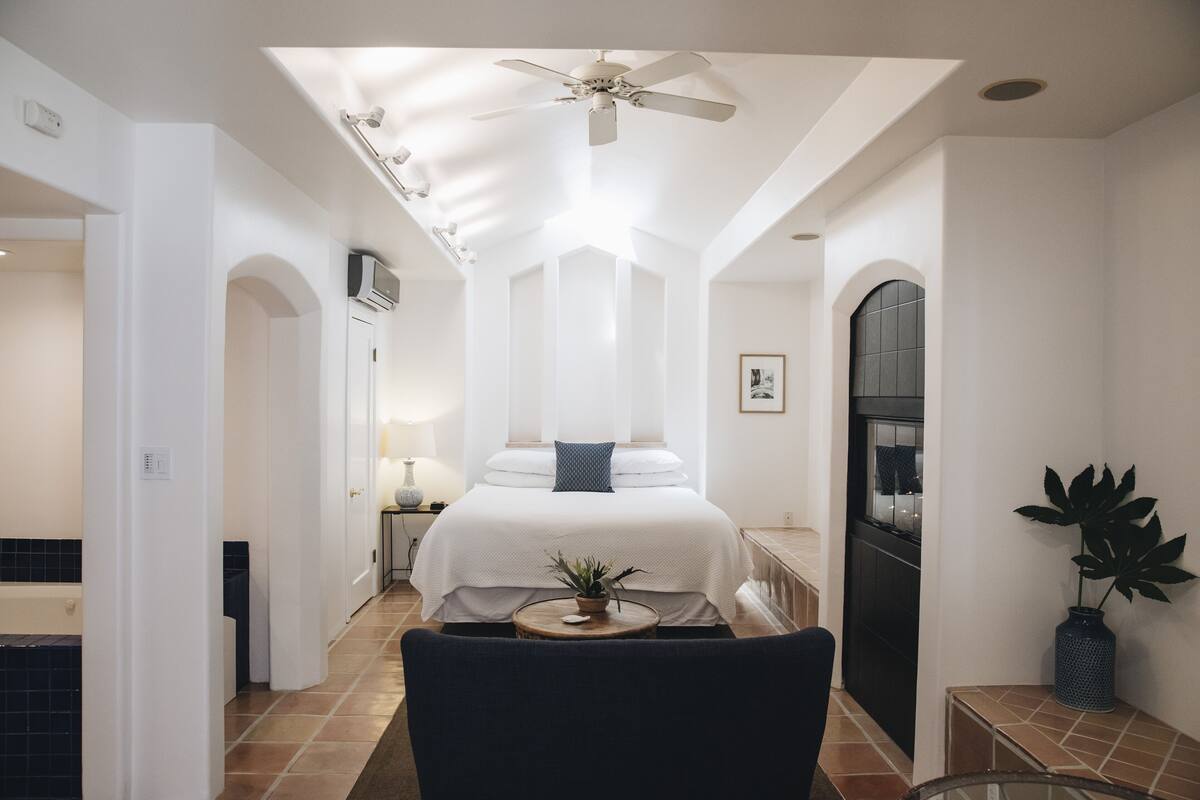 A bedroom with a bed, a ceiling fan, a nightstand, a lamp, a potted plant, and a view of the kitchen.
