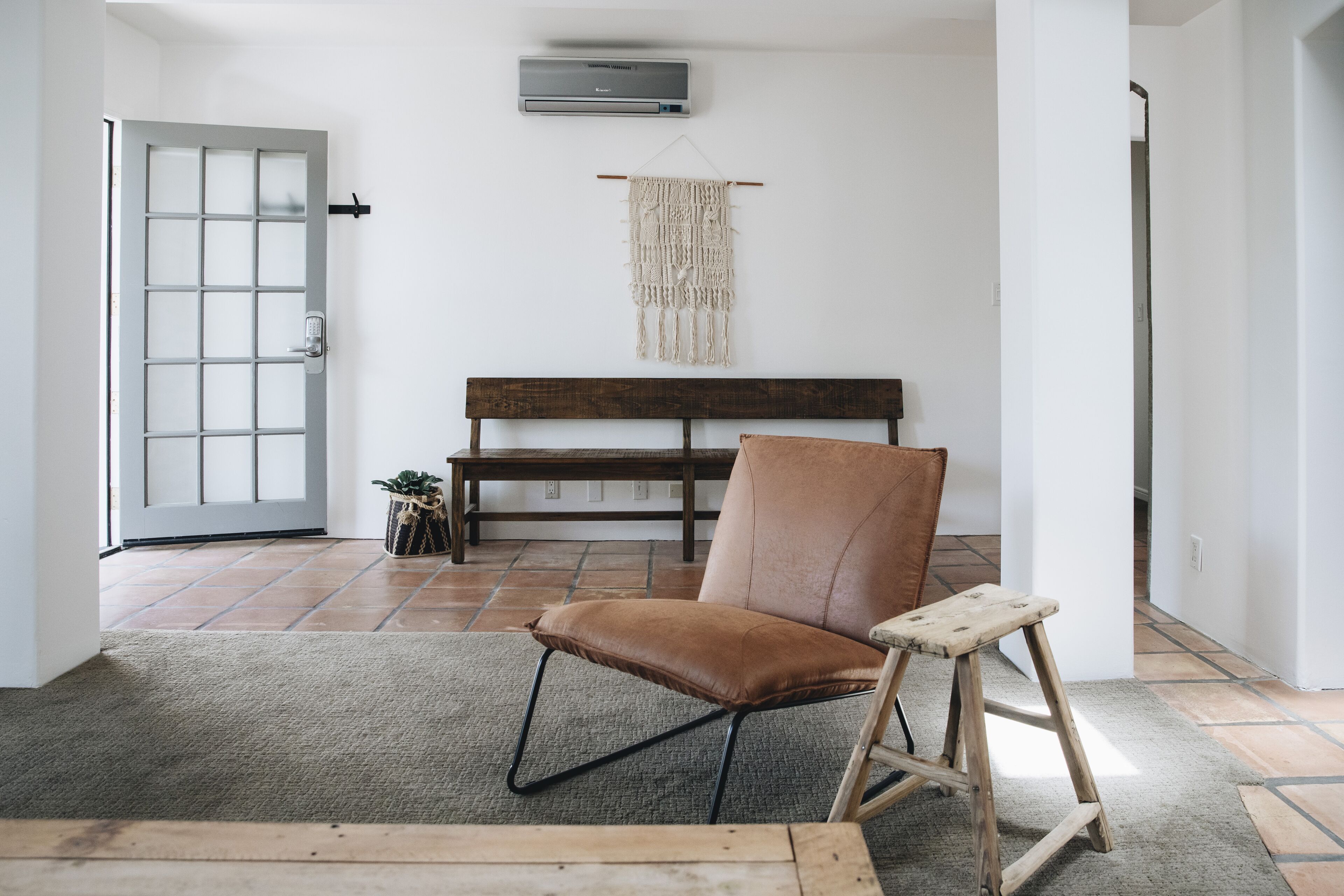 A room with a wooden bench, a brown leather chair, a small wooden stool, a macrame wall hanging, and a door with a frosted glass panel.