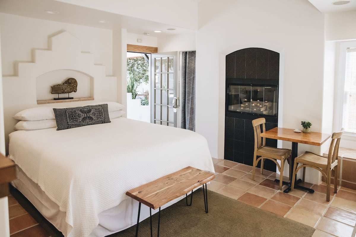 A bedroom with a large bed, a wooden bench, a dining table with chairs, and a fireplace.