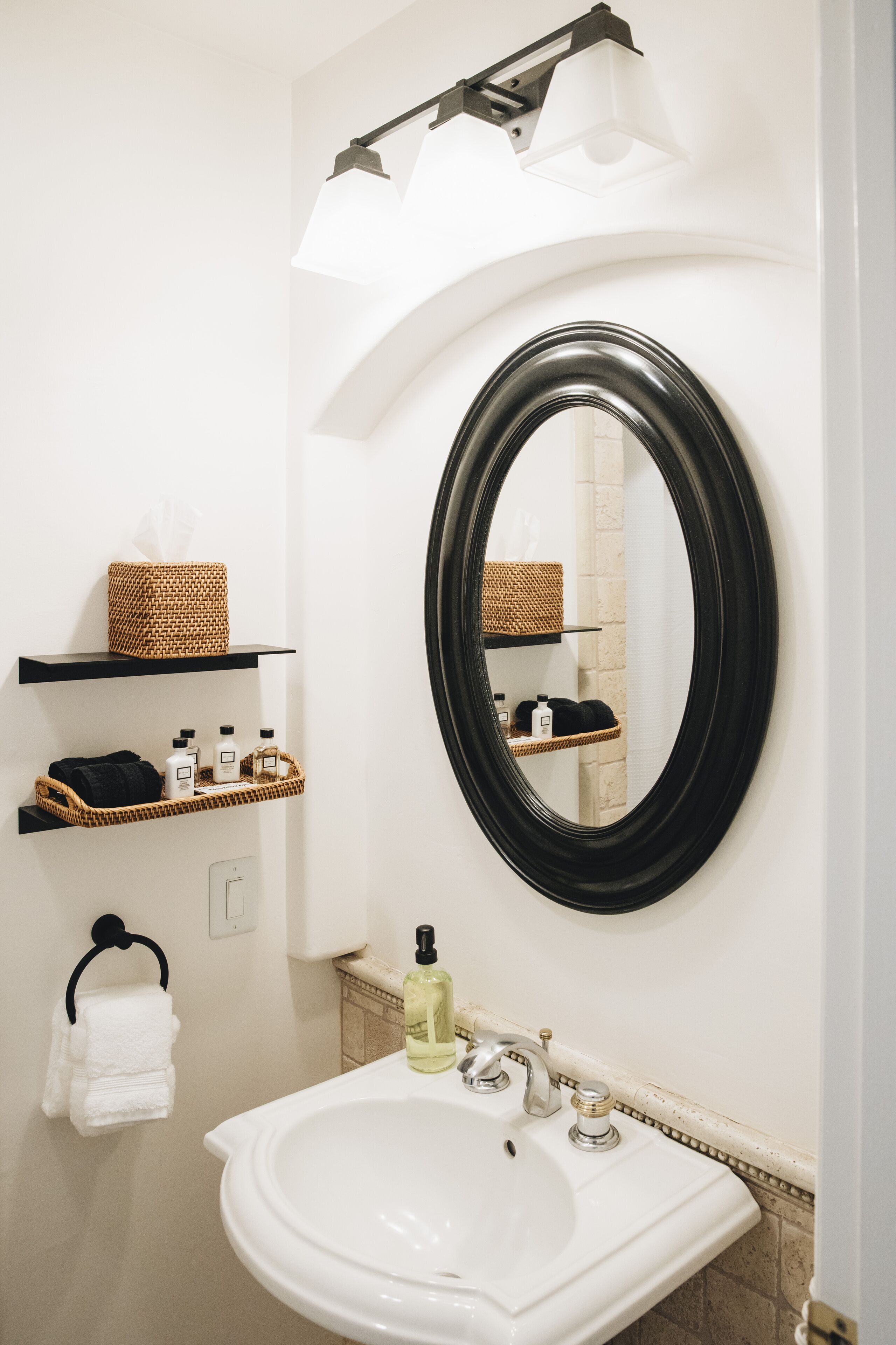 Un baño con un lavabo blanco, un espejo con marco negro y estantes con artículos de aseo.