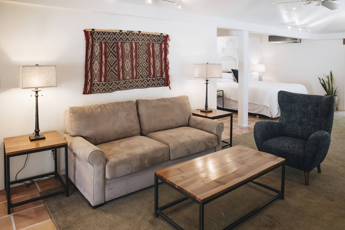 A living room with a beige sofa, a wooden coffee table, a dark armchair, and a patterned throw on the wall.