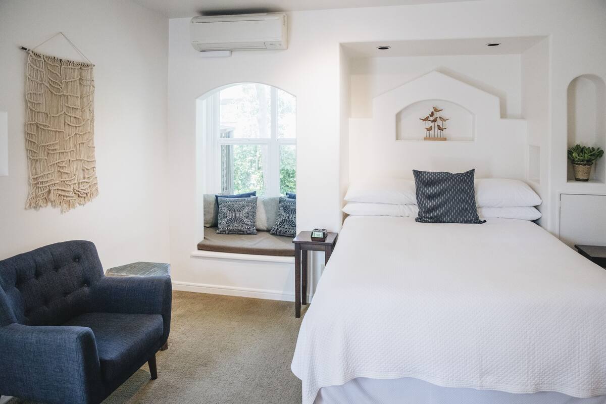 A bedroom with a large bed, a window seat, a dark blue armchair, and a macrame wall hanging.