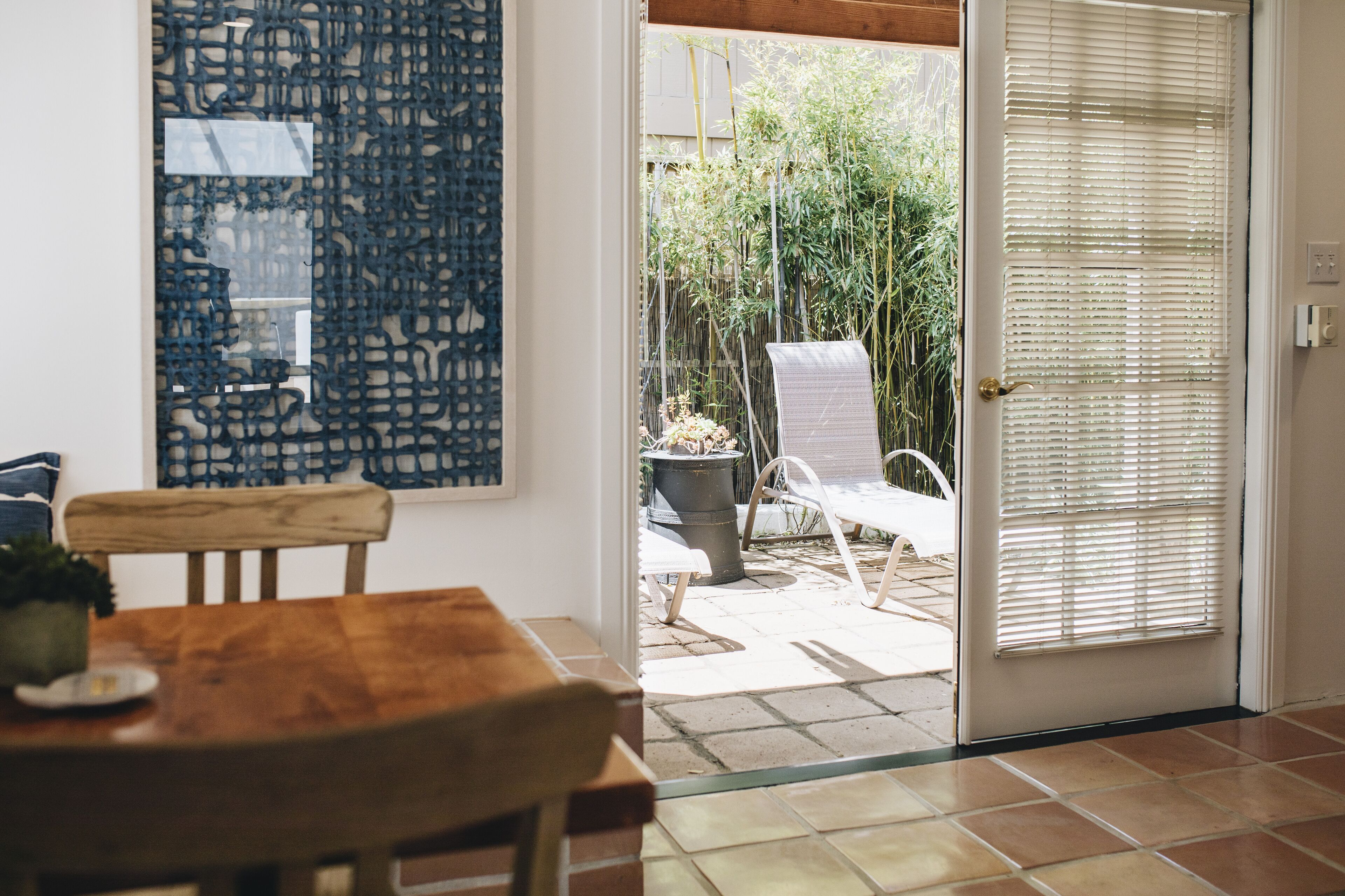 Una mesa de comedor de madera con sillas, un cuadro abstracto grande en la pared y un ventanal corredero que da a un patio exterior con una silla y un bote de basura.