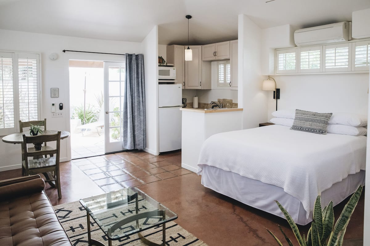 A modern hotel room with a large bed, a small kitchenette, a dining area, and a view of the outdoors.