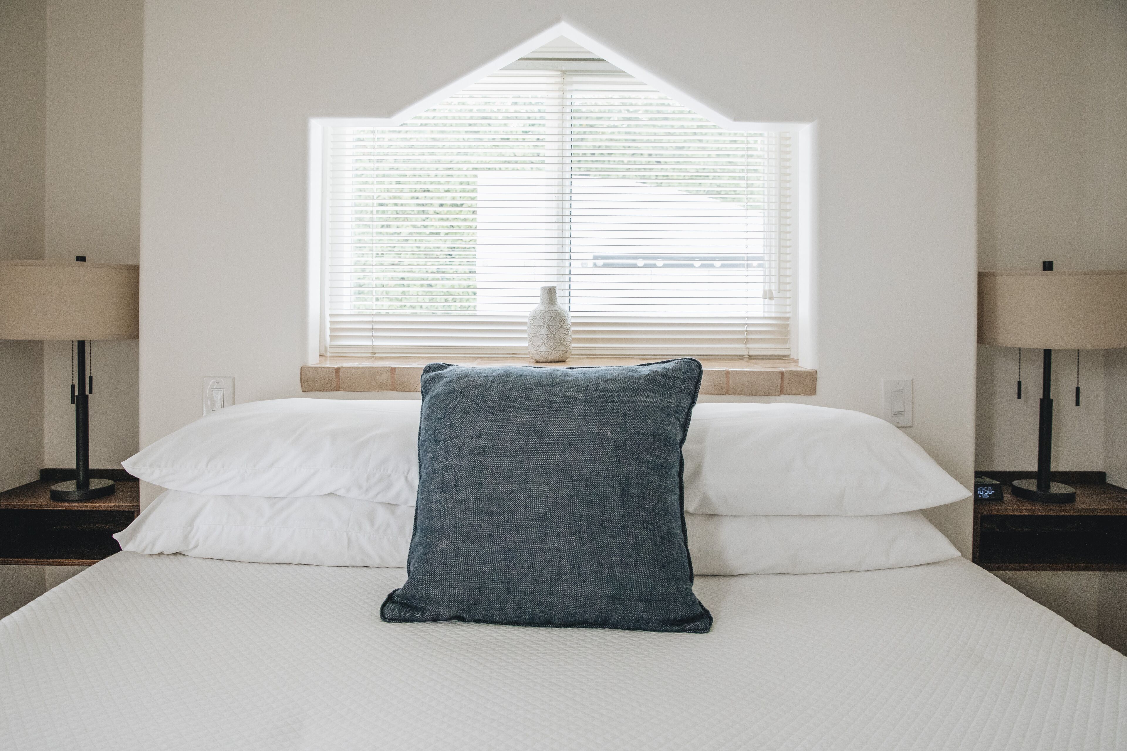 A bedroom with a bed, white pillows, a blue pillow, a nightstand, a lamp, and a window with blinds.