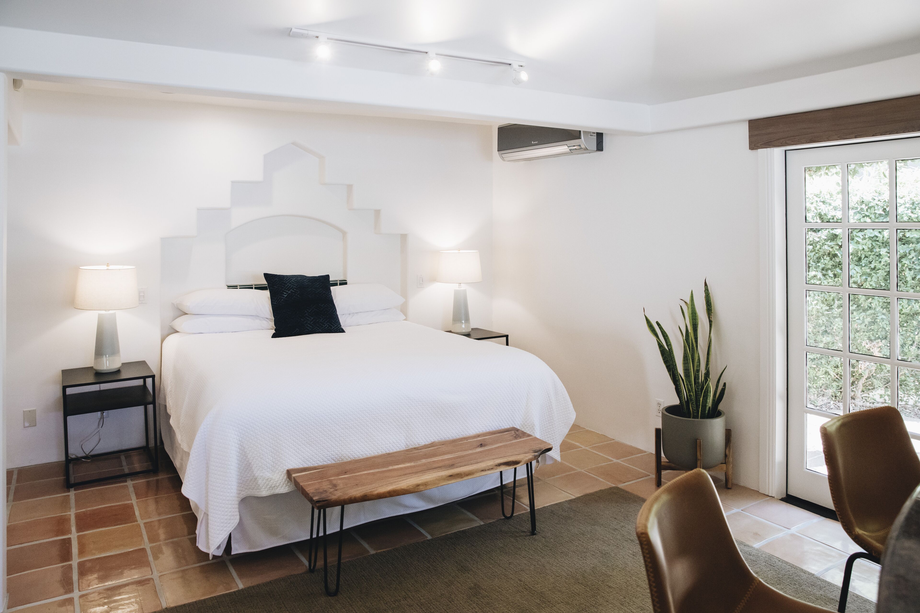 Un dormitorio con una cama grande, dos mesitas de noche con lámparas, un banco, una planta en maceta y vistas a la vegetación a través de una ventana.