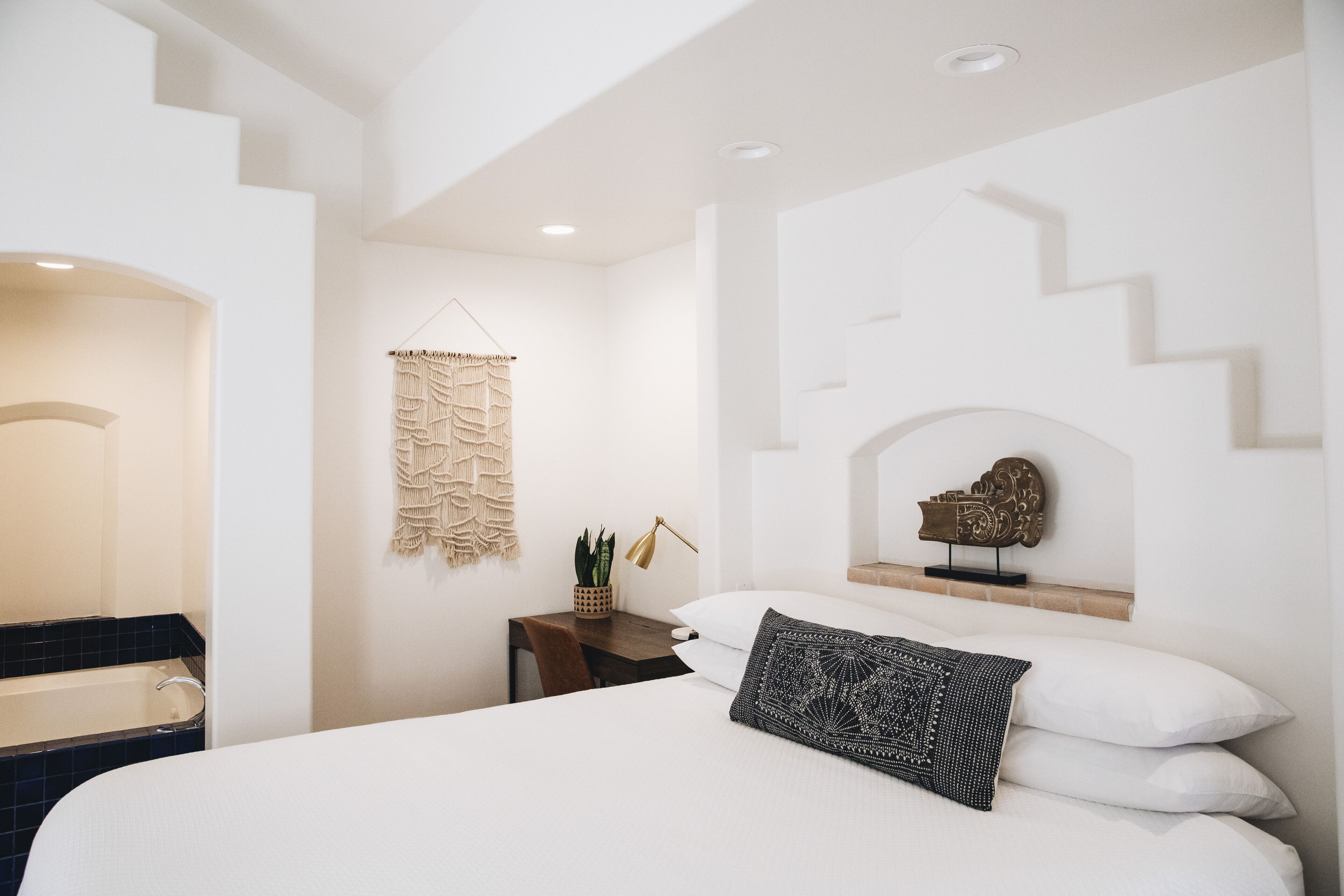 Un dormitorio moderno con una cama grande, un escritorio pequeño, un macramé colgado en la pared y un adorno decorativo en la pared.