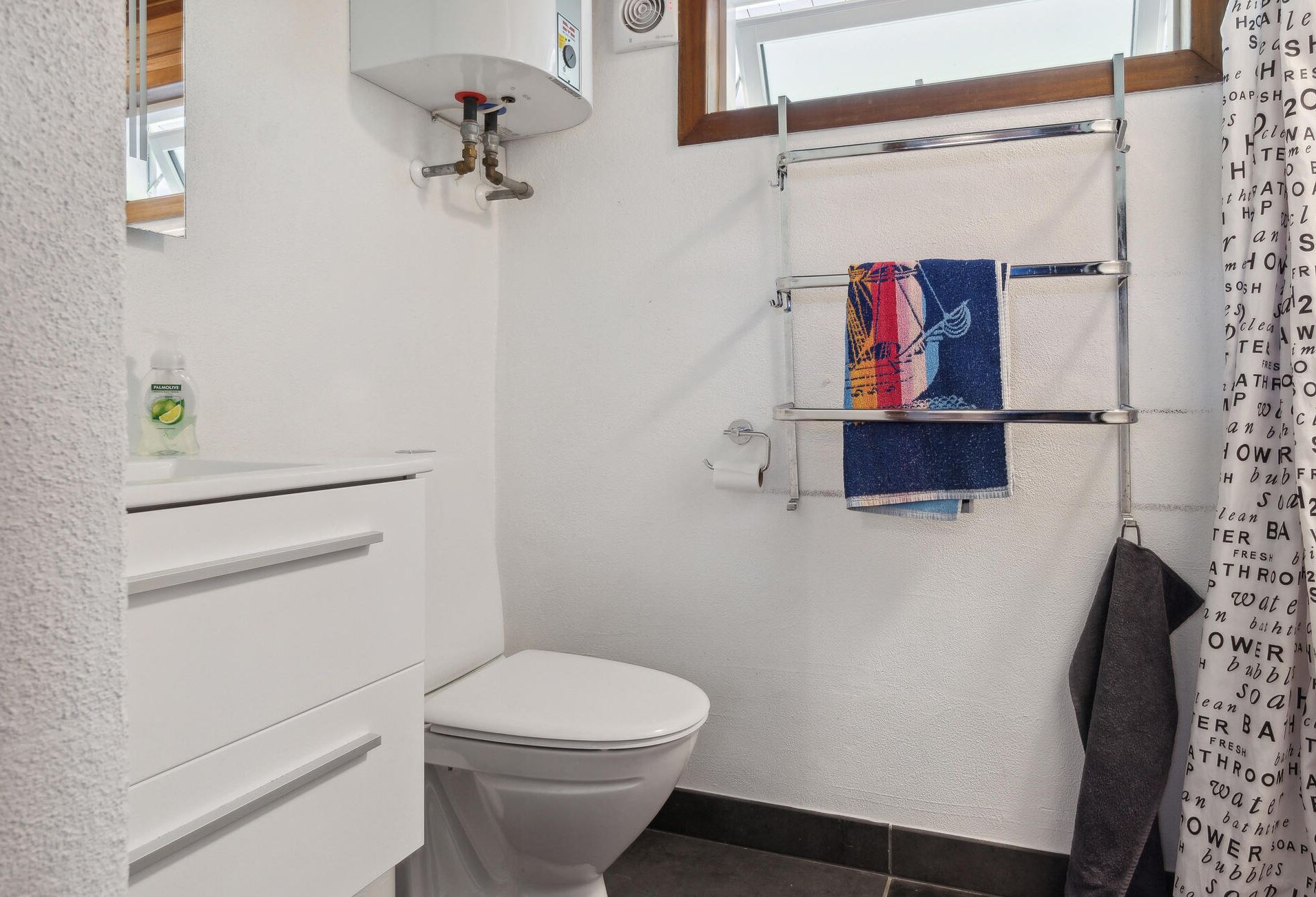Un baño con inodoro, toallero con una toalla colorida, una botella de jabón líquido y cortina de ducha.