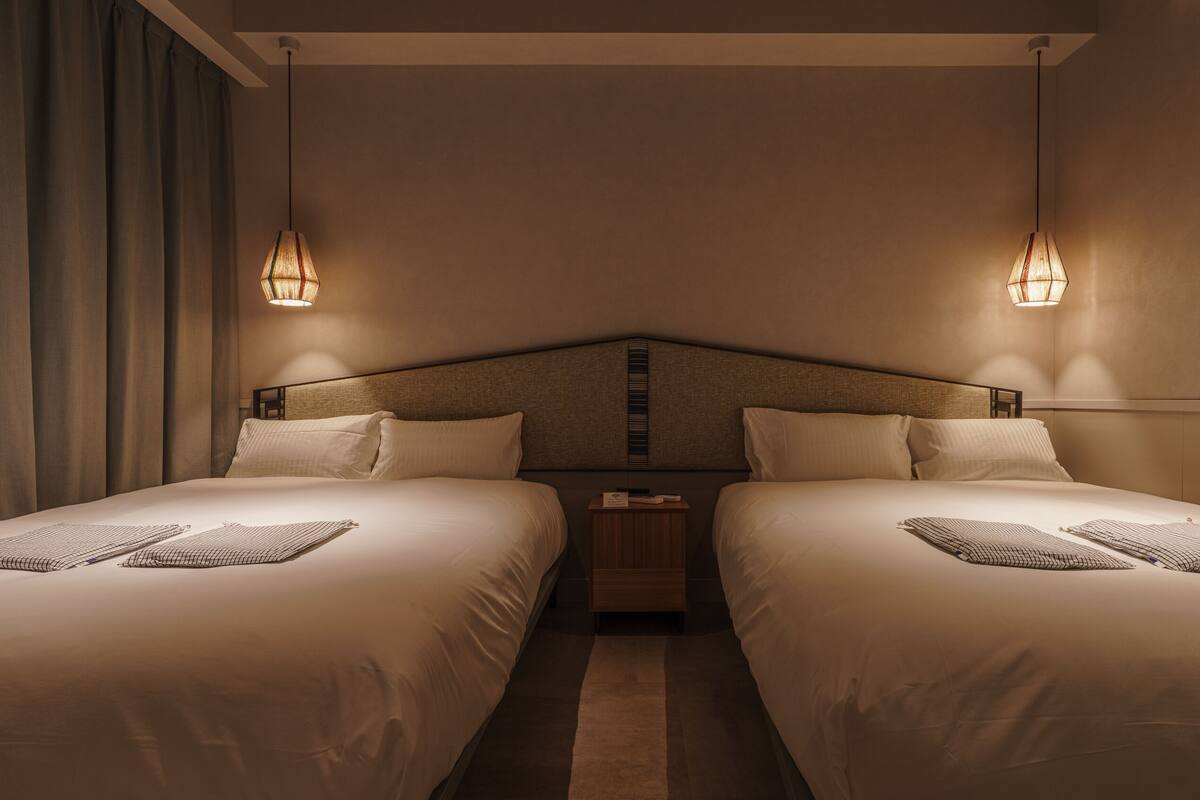 A hotel room with two single beds, a nightstand, and two wall-mounted lamps.