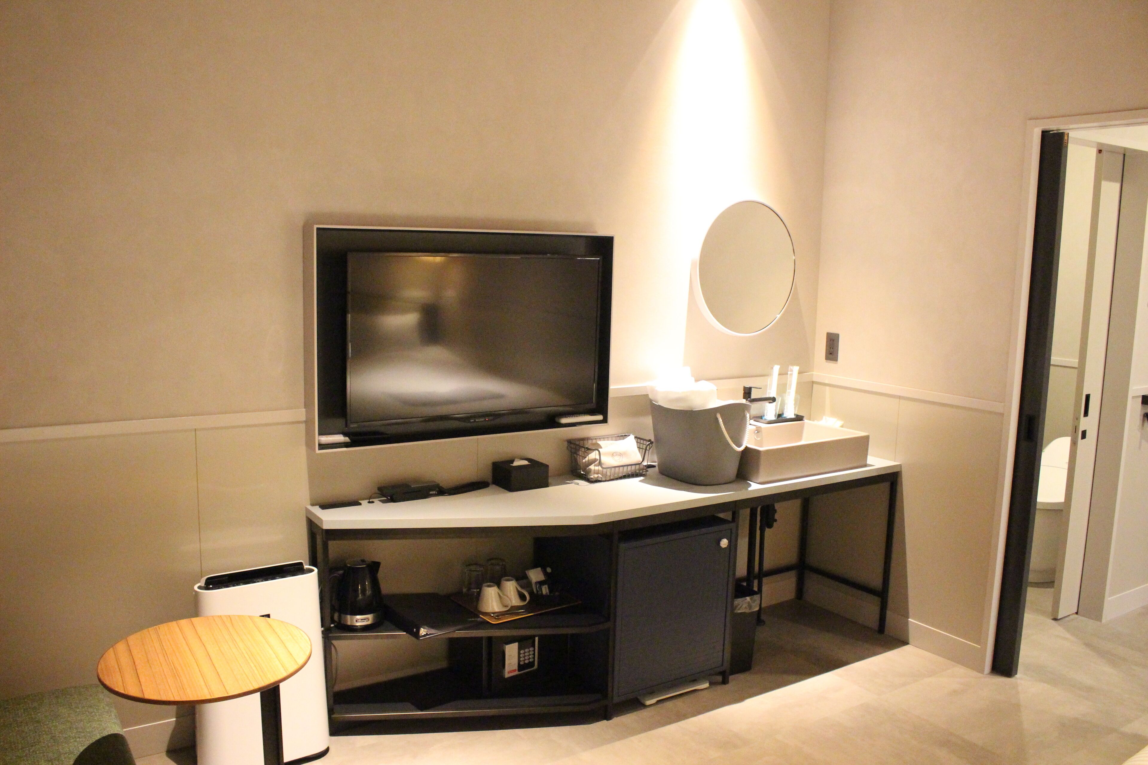 Une chambre d’hôtel moderne équipée d’une télévision à écran plat, d’un petit bureau avec un miroir, d’une machine à café et d’une poubelle.