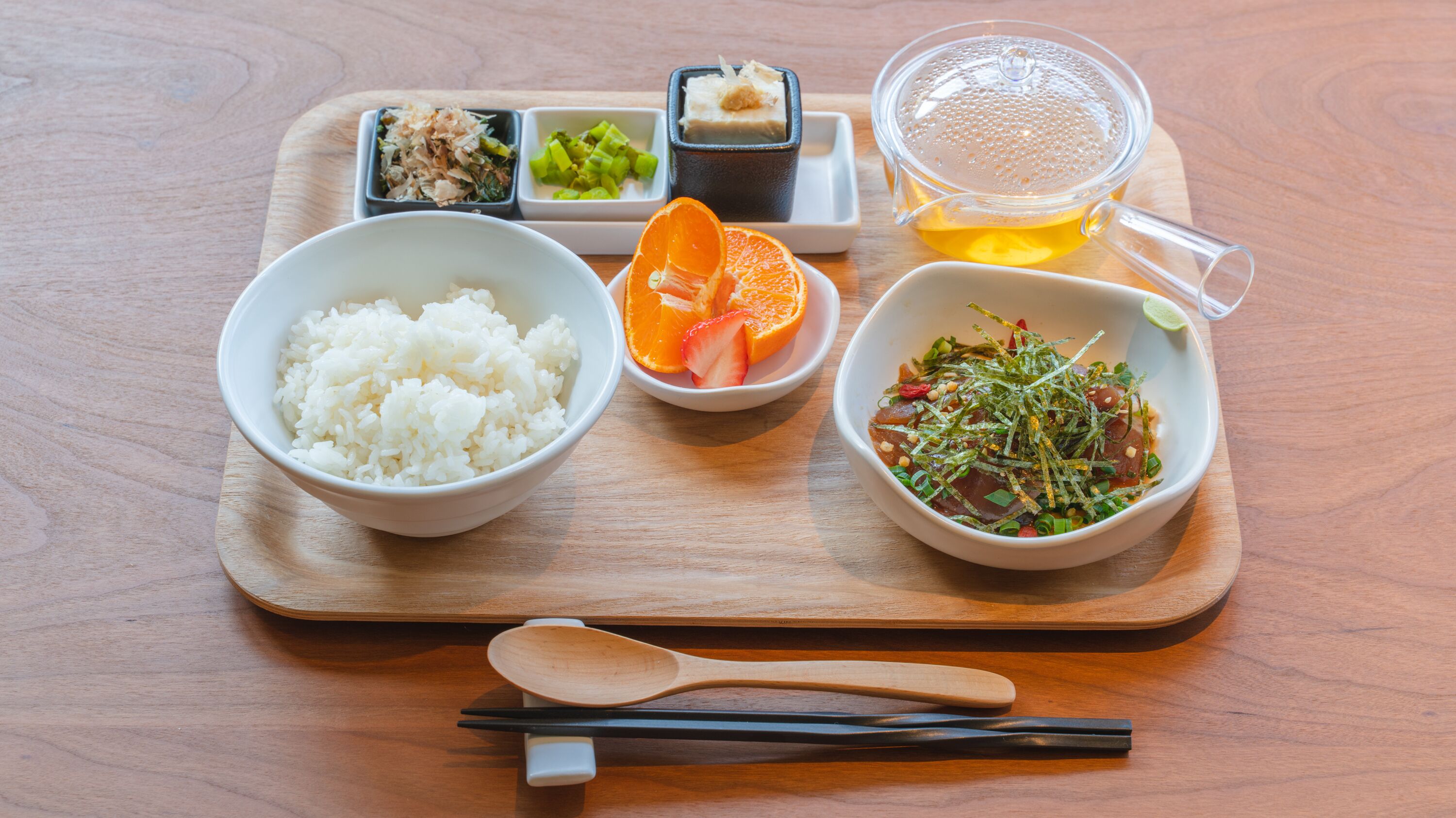 Un plateau en bois avec des bols de riz, des tranches d’orange, une salade et un verre de thé.