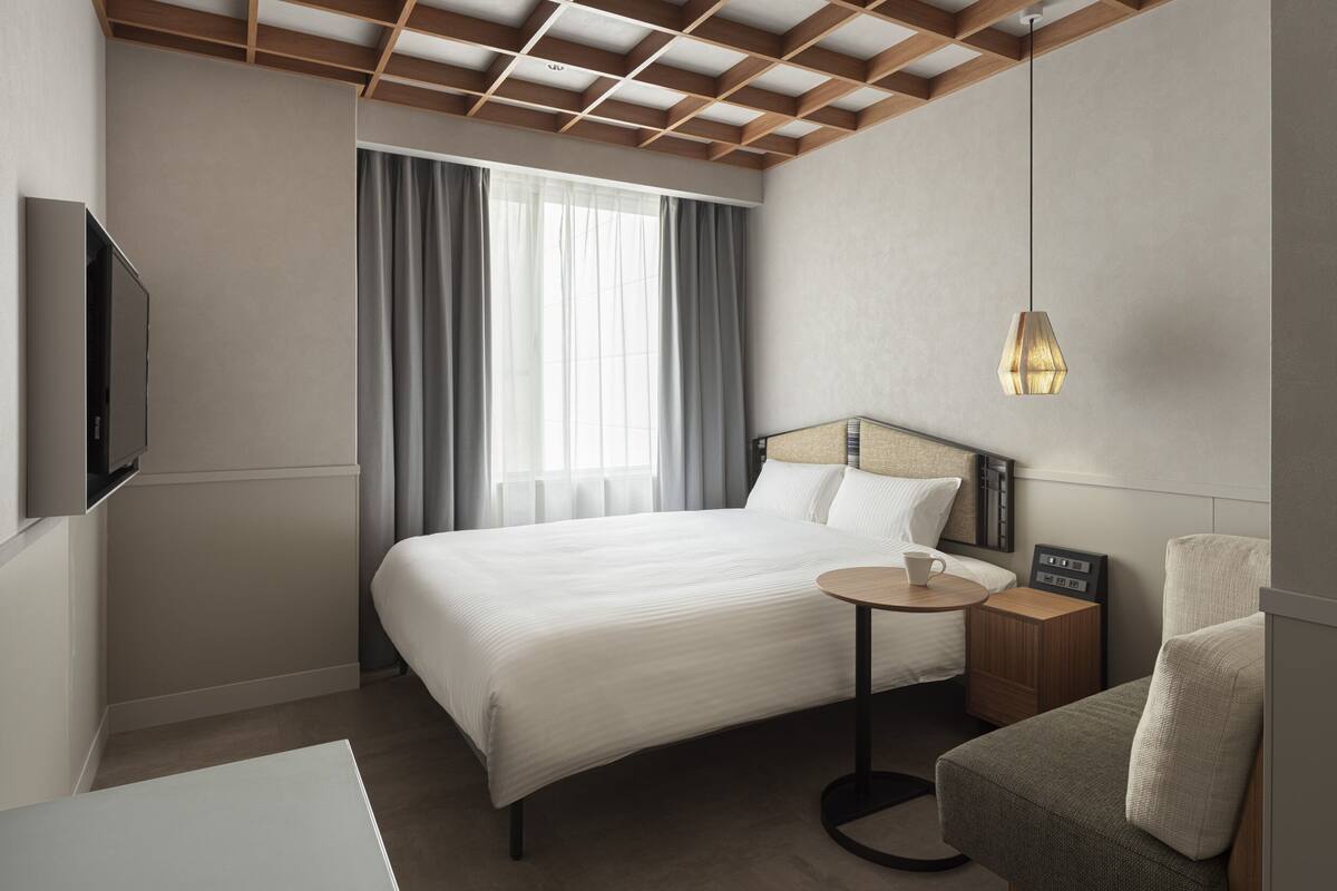 A modern hotel room with a large bed, a bedside table with a cup and a radio, a TV mounted on the wall, and a hanging pendant light.