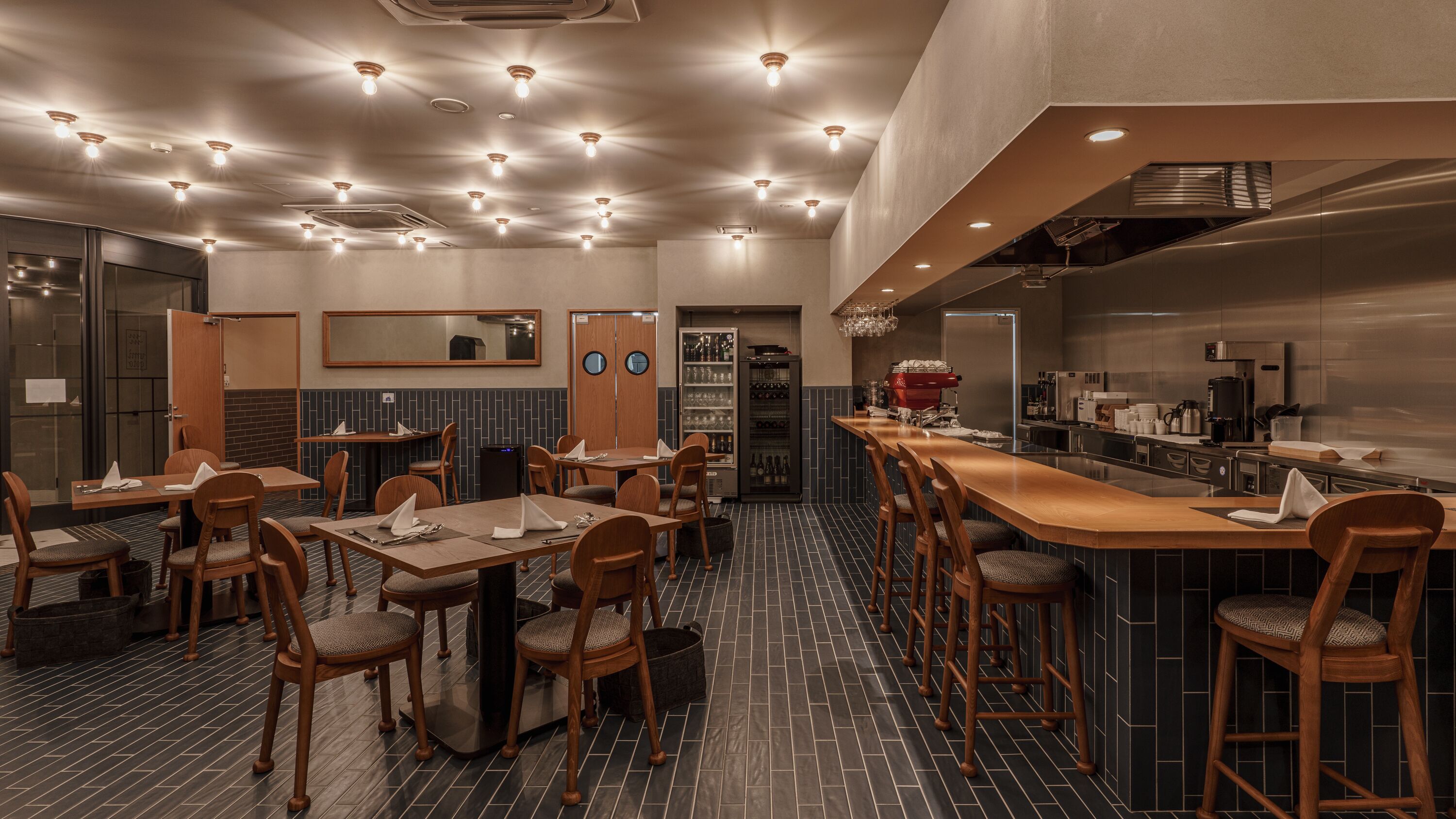 Ein modernes Restaurant mit mehreren Tischen, Stühlen, einer Bar und einer im Hintergrund sichtbaren Küche.