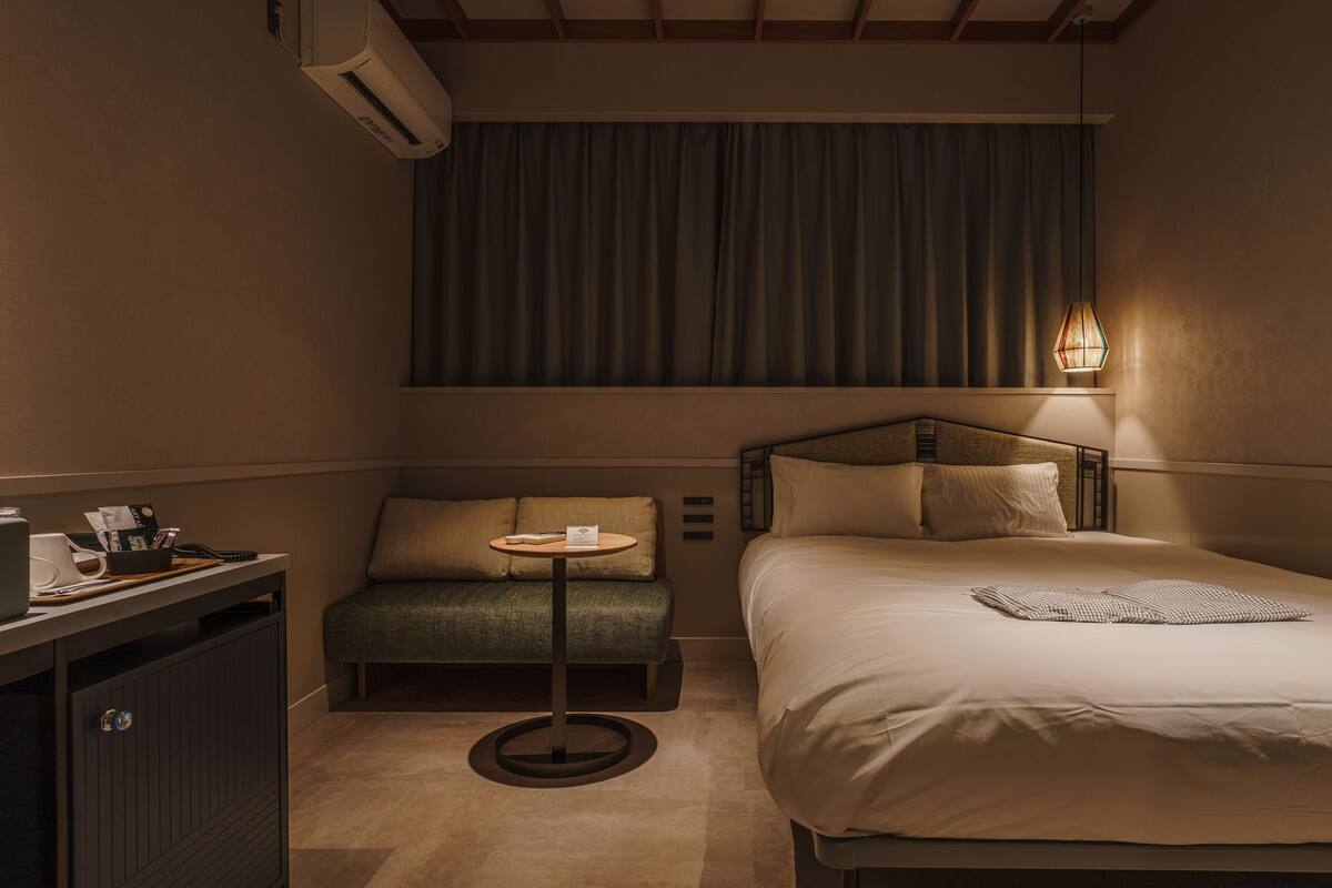 A compact hotel room with a single bed, a small desk with a telephone and papers, a chair, and a bedside table.