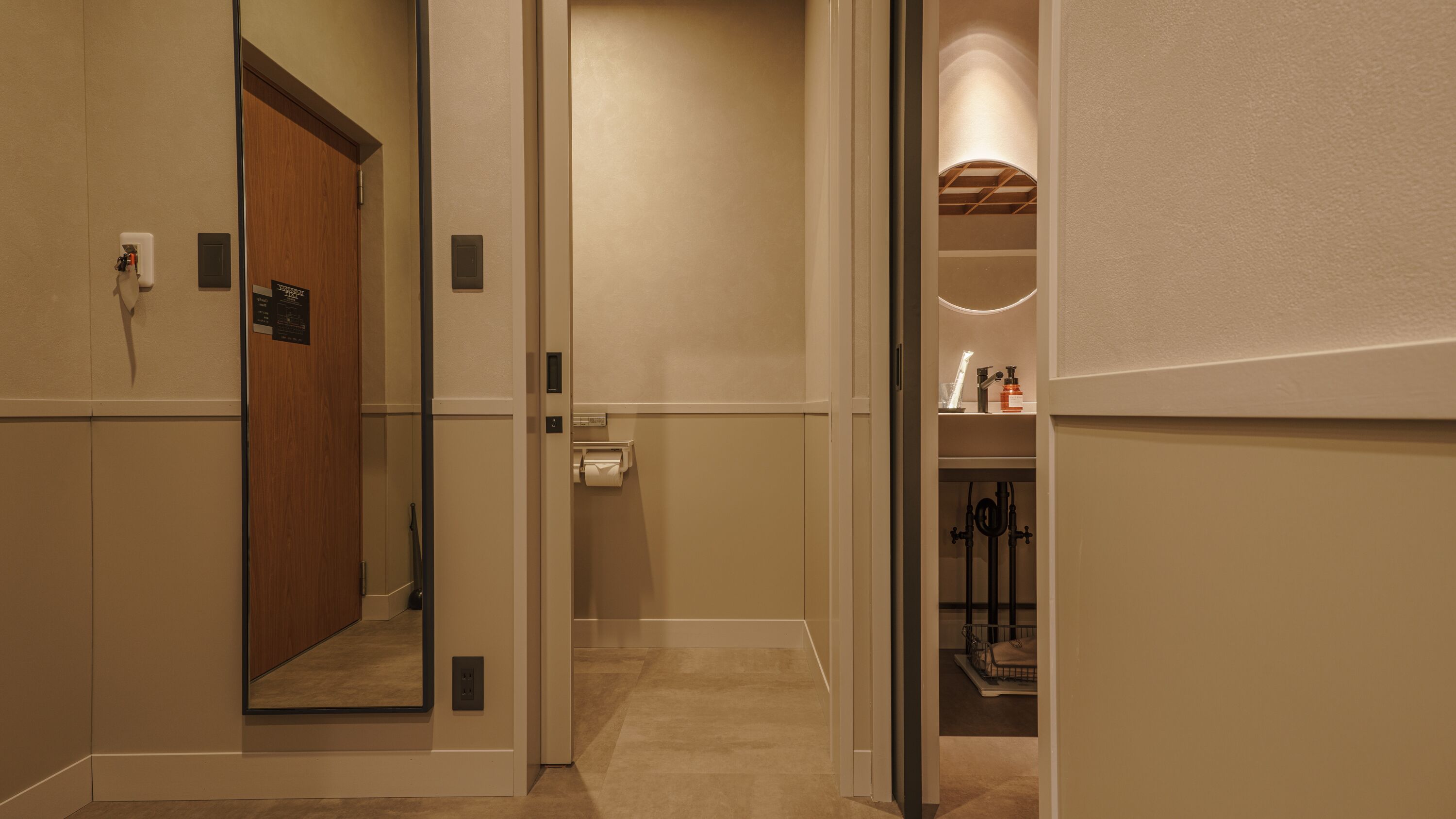 A hotel hallway with a wooden door, a hand dryer, a paper towel dispenser, and a small shelf with toiletries.