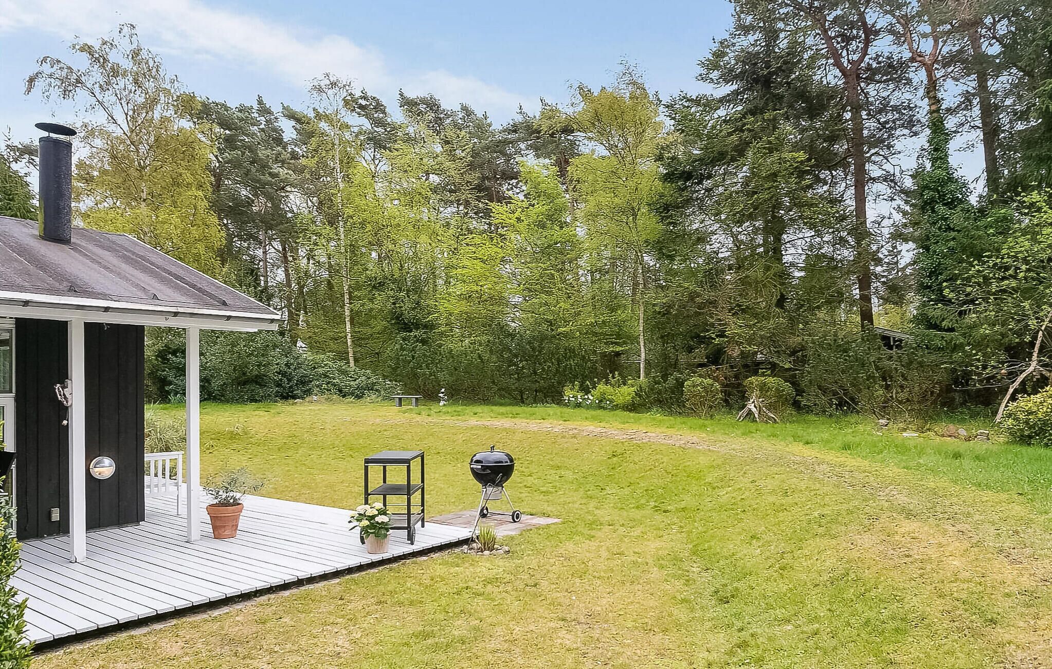 A wooden deck with a grill and potted plants, surrounded by a grassy yard and dense trees.