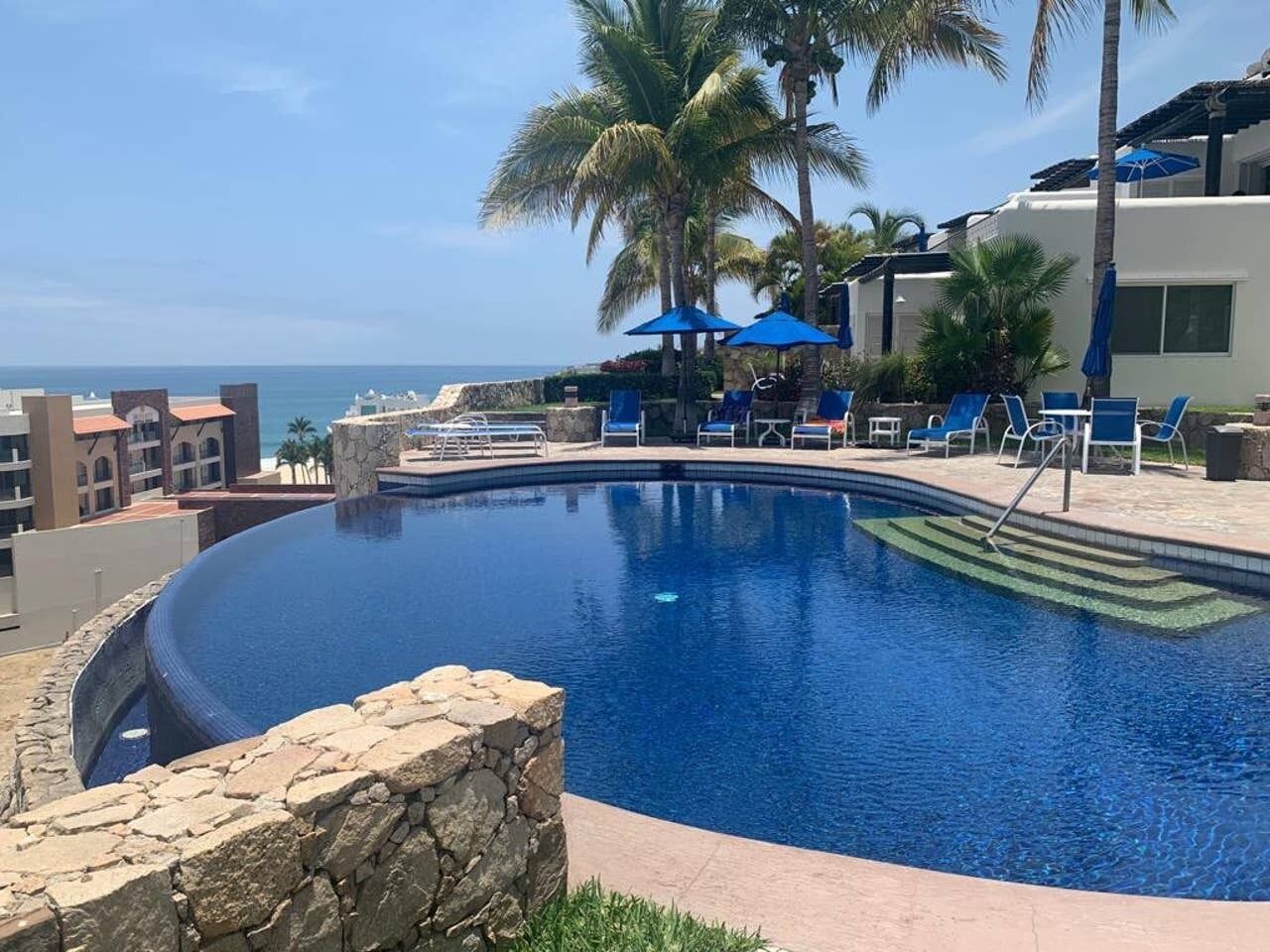 A pool with lounge chairs and umbrellas, palm trees, and a view of the ocean.