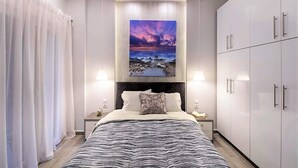 A modern bedroom with a bed, nightstands, a headboard, a wardrobe, and a painting of a sunset over a rocky beach. - Thessaloniki Historic Center - Metro Apartment (Thessaloniki)