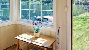 Una habitación con una mesa de madera, una ventana con vistas a un lago y un barco, y una planta pequeña en maceta.