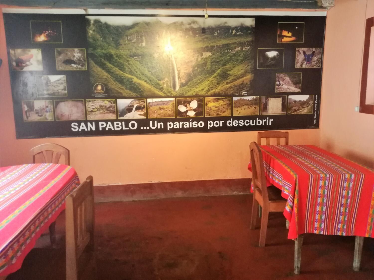A room with two wooden tables, each covered with a red tablecloth. A large poster on the wall displays various photographs of San Pablo, including a waterfall and a mountain landscape.