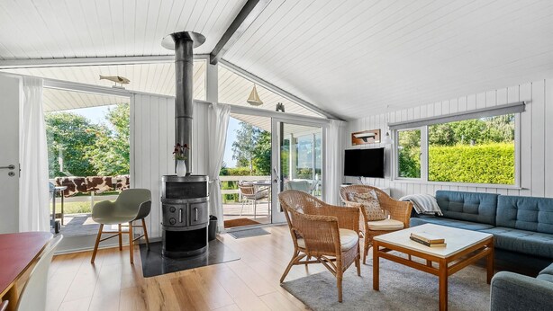 A spacious living room with a wood-burning stove, wicker furniture, a blue sofa, and large windows offering a view of the outdoors. - Cozy home in Nysted with house sea view (Nysted)