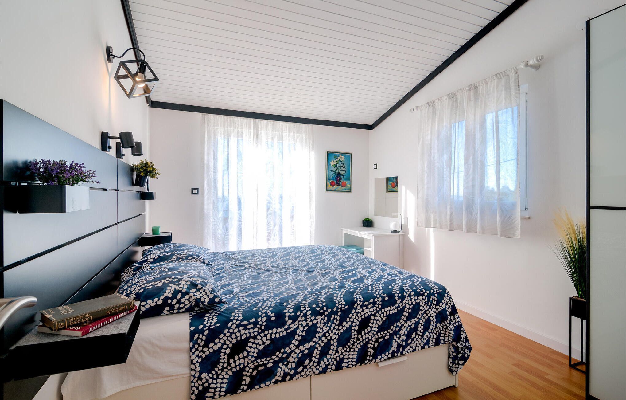 Een slaapkamer met een bed, nachtkastjes, een bureau, een stoel, een hanglamp en een raam met dunne gordijnen.