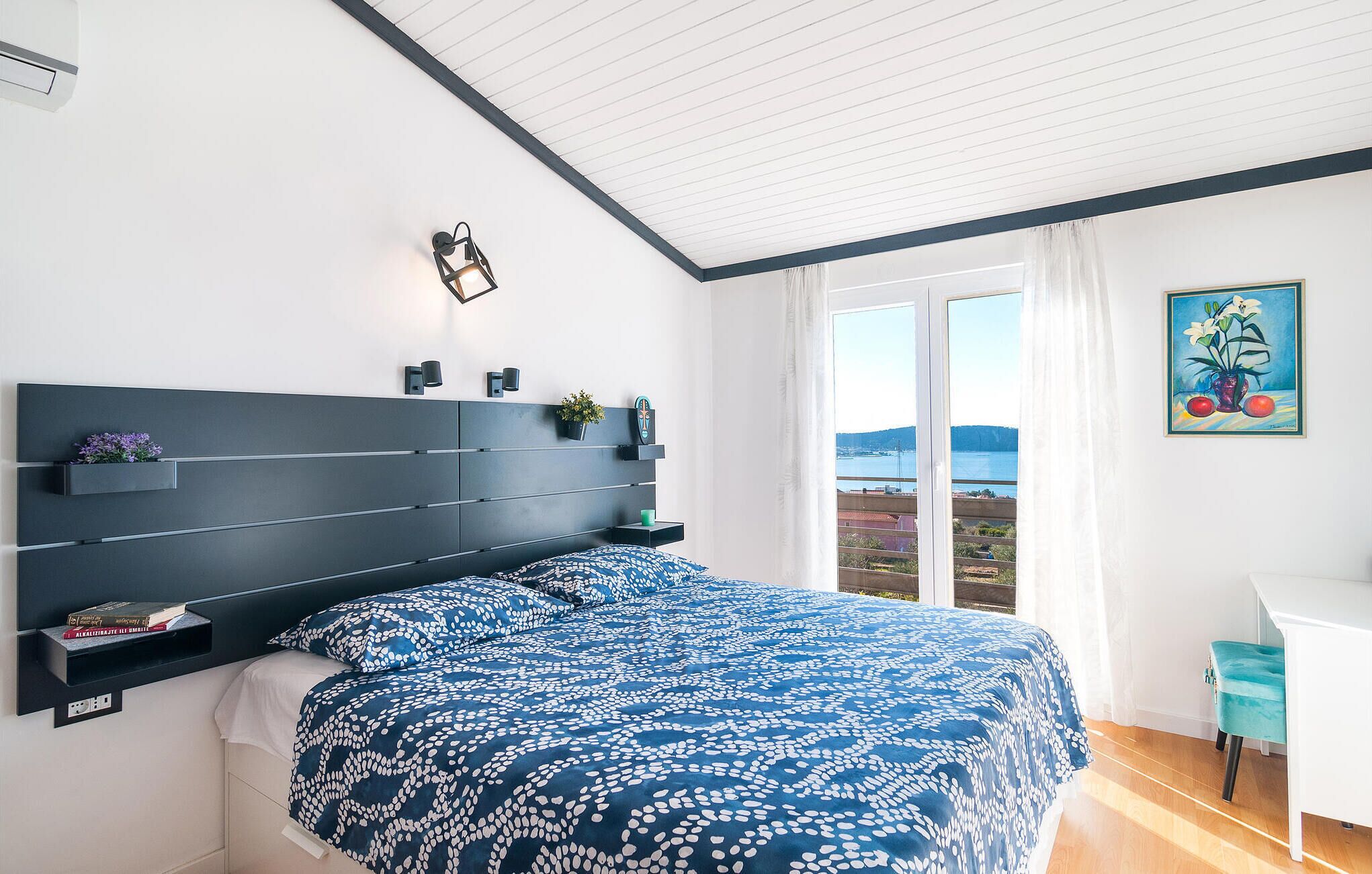 Ein Schlafzimmer mit einem großen Bett, einem Kopfteil mit integrierten Regalen, einem Fenster mit Meerblick und einem Gemälde an der Wand.