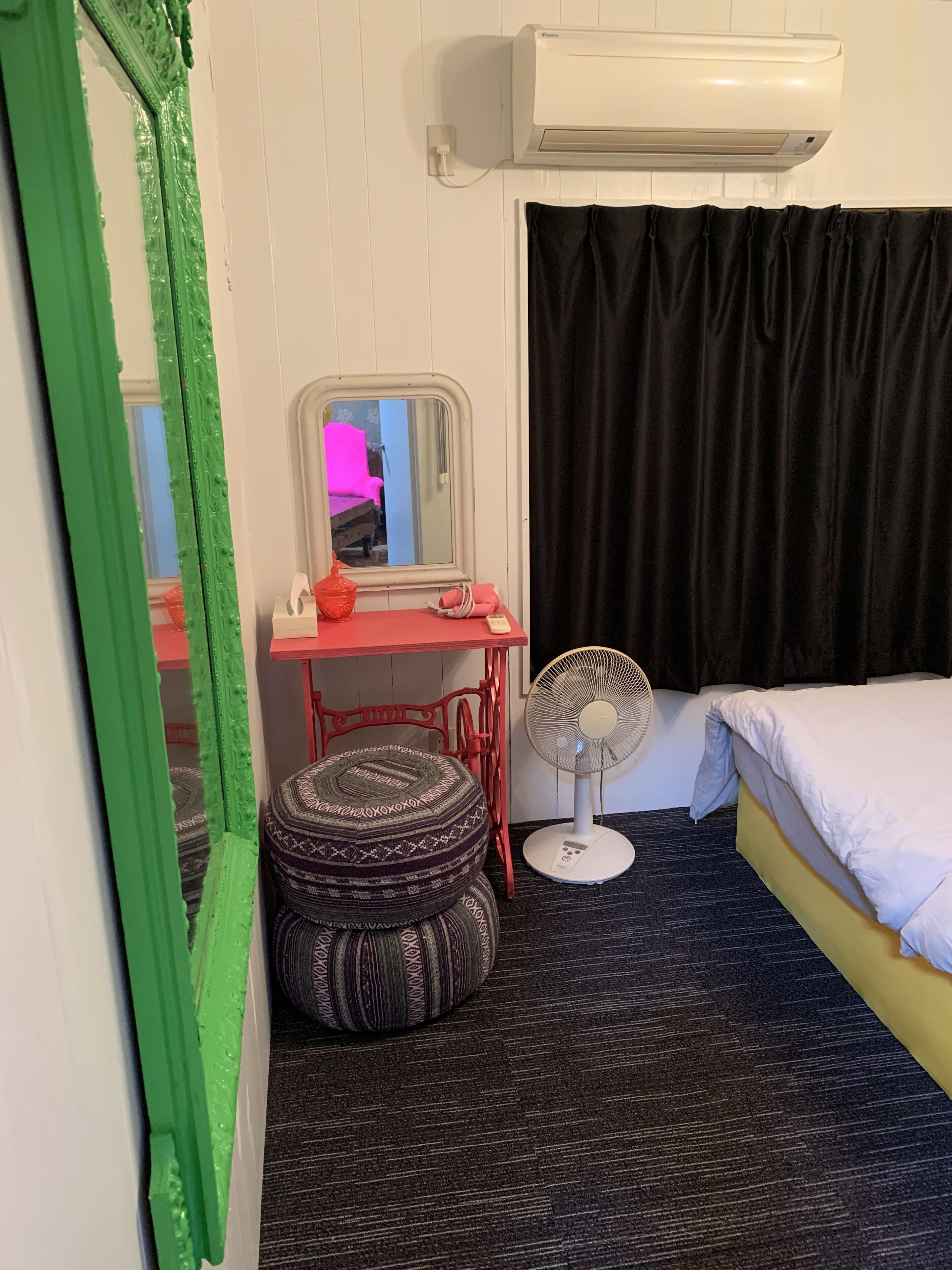 Una habitación de hotel con una cama, una mesita de noche con un espejo, un ventilador de pie y un reposapiés estampado.