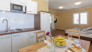 Une cuisine moderne avec des meubles blancs, un four à micro-ondes et un évier. On y trouve également un coin repas avec une table en bois dressée pour le petit-déjeuner, comprenant des fruits, des viennoiseries et un vase avec une fleur.