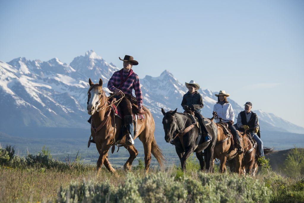 A group of people riding horses on a grassy field with a mountainous backdrop.