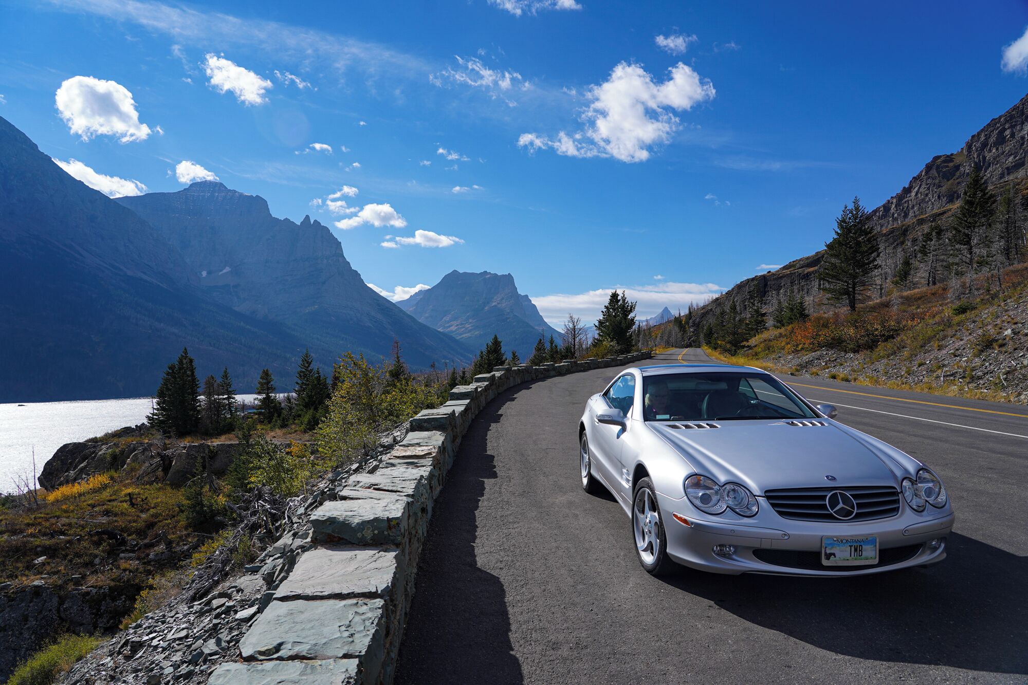 A silver car parked on a paved road with a stone guardrail, surrounded by mountains and trees.