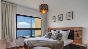 A modern bedroom with a large window, a wooden headboard, a bedside table, and a hanging light fixture. - Centromar Bella Vista by Atlantic Holiday (Funchal)