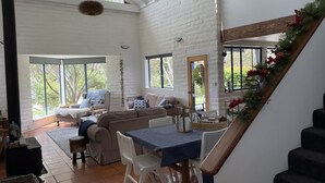 A spacious living area with a large window, a sofa, a dining table, and a staircase. - Lakefront@Tyrolean House Waterfront EAST JINDABYNE (East Jindabyne)