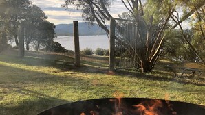 A campfire in a metal fire pit with logs and burning wood, surrounded by grass and trees. - Lakefront@Tyrolean House Waterfront EAST JINDABYNE (East Jindabyne)