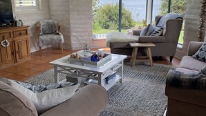 A modern living room with a large window offering a lake view, a cozy seating area, and a flat-screen TV on a wooden cabinet. - Lakefront@Tyrolean House Waterfront EAST JINDABYNE (East Jindabyne)