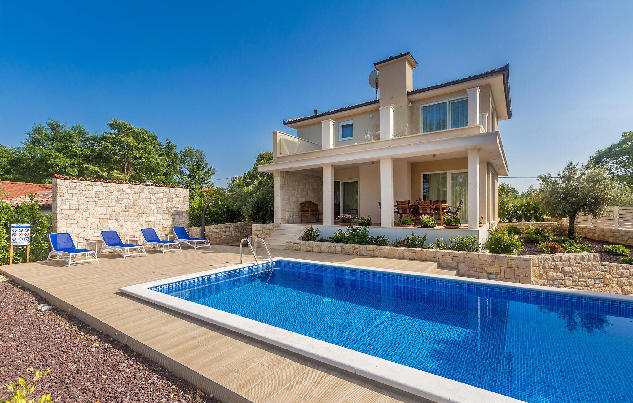 A two-story house with a swimming pool, lounge chairs, and a stone wall.