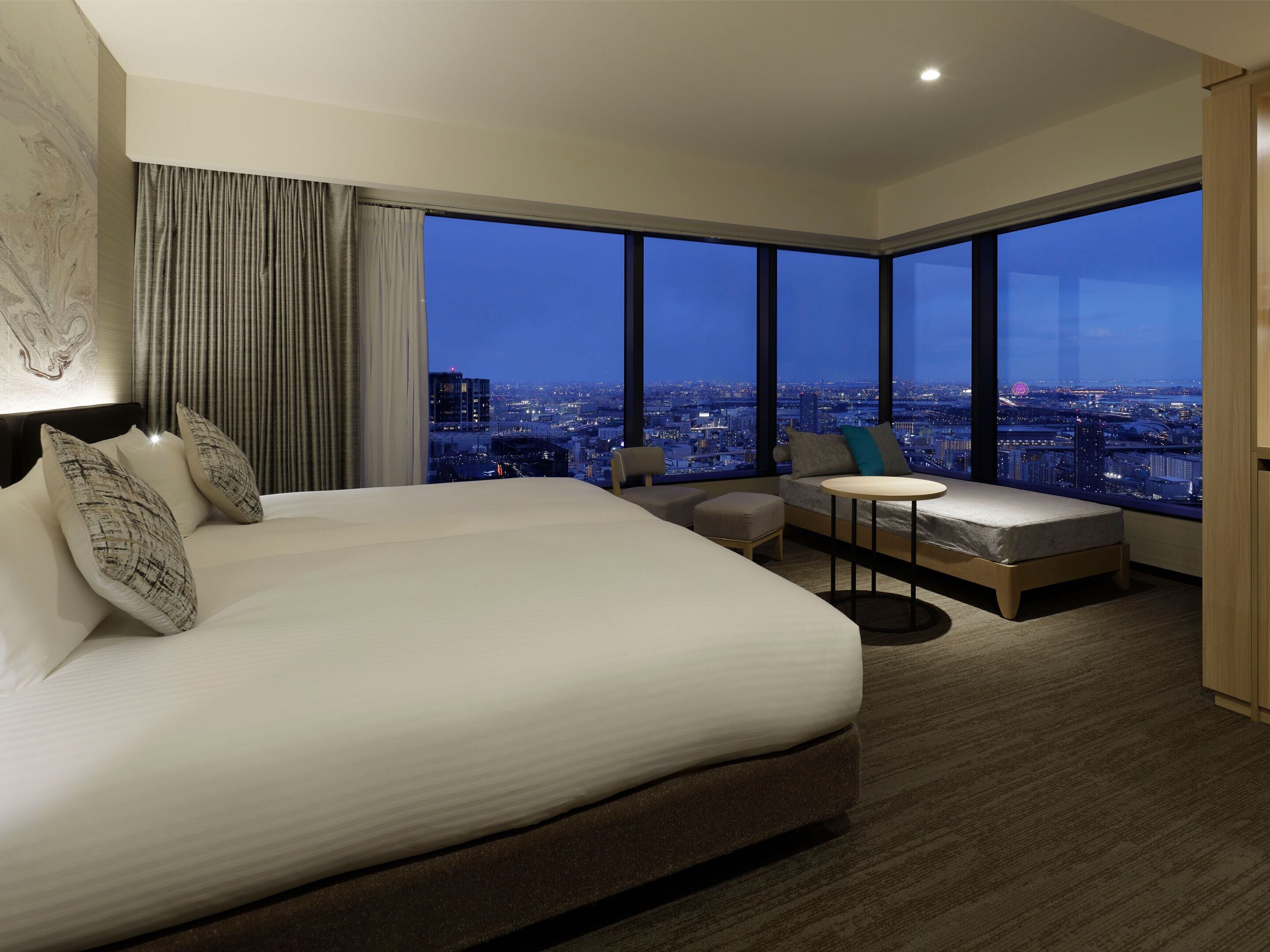 Een hotelkamer met een groot bed, een kleine zithoek, een ronde tafel en een stadszicht door grote ramen.