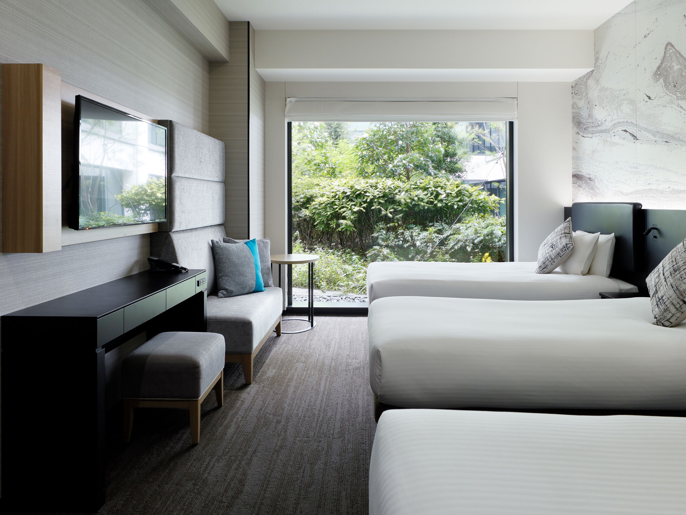 A modern hotel room with a bed, a desk, a chair, and a view of greenery outside.