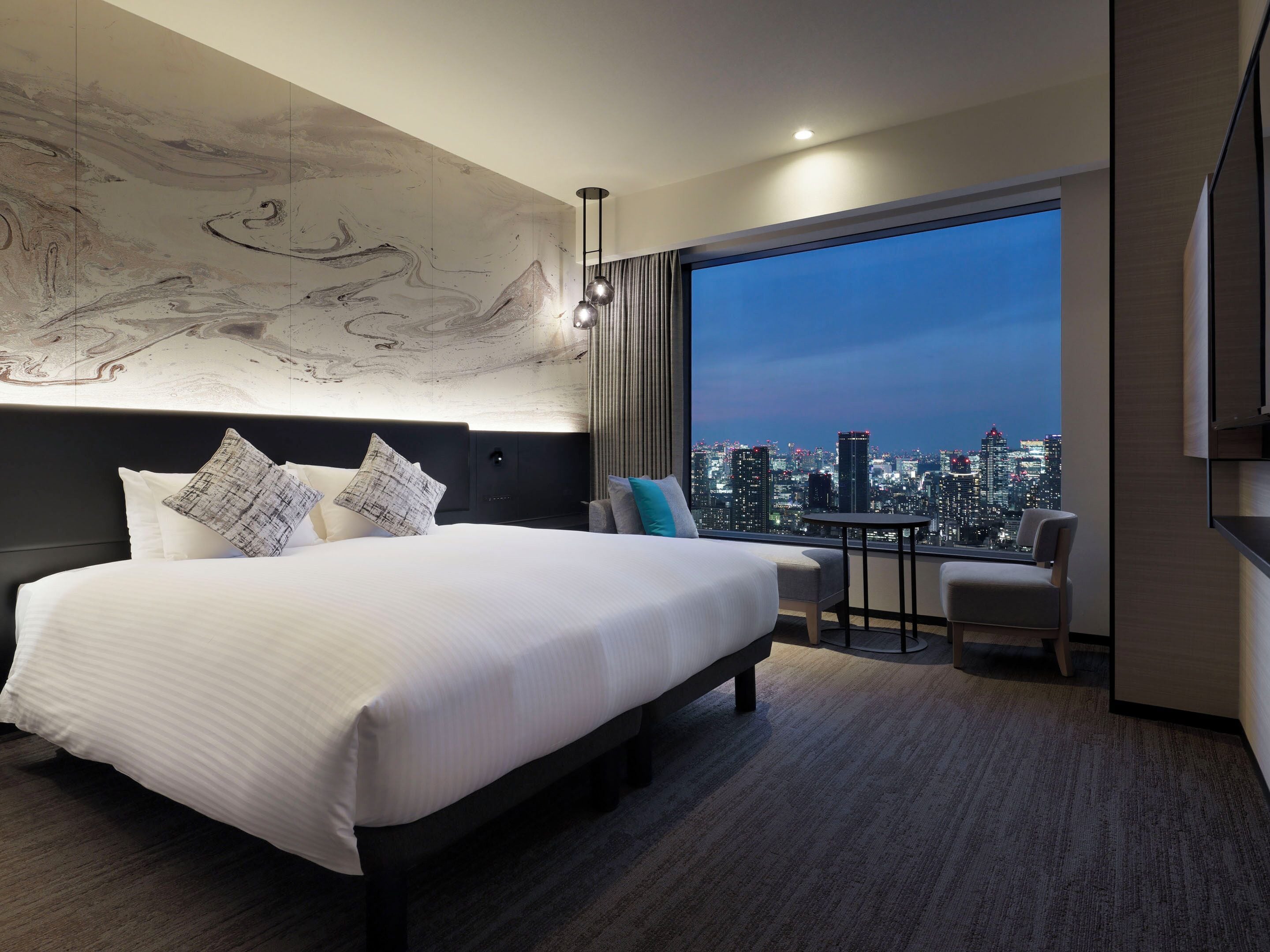 A modern hotel room with a large bed, a city view, a small table, and a chair.