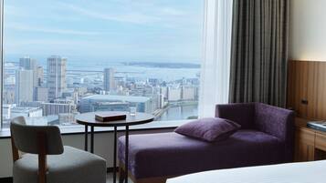 Une chambre d’hôtel avec vue sur la ville, une table ronde avec un livre et un verre, un canapé violet et un lit.