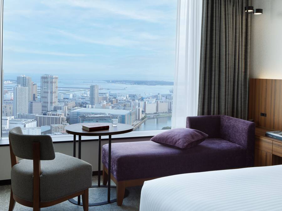 Une chambre d’hôtel avec vue sur la ville, une table ronde avec un livre et un verre, un canapé violet et un lit.