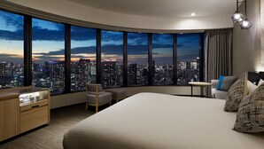 Habitación de hotel moderna con una cama grande, un escritorio, una silla, una mesita y vistas a un paisaje urbano nocturno.