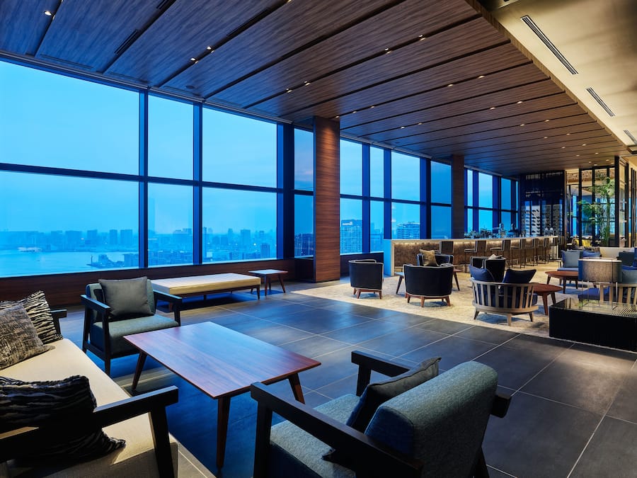 A spacious lounge area with large windows offering a city view, a bar area with seating, and a mix of modern and classic furniture.