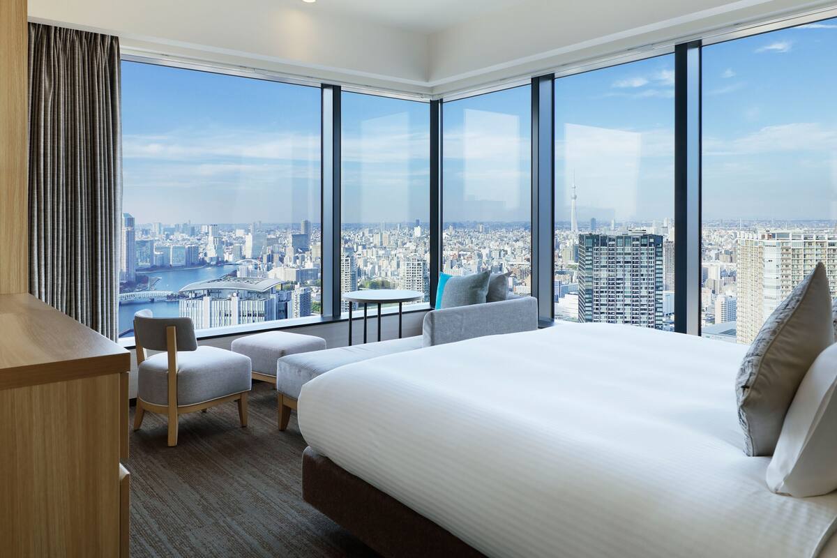 Habitación de hotel moderna con una cama grande, una mesita pequeña, una silla y vistas a un paisaje urbano a través de amplias ventanas.