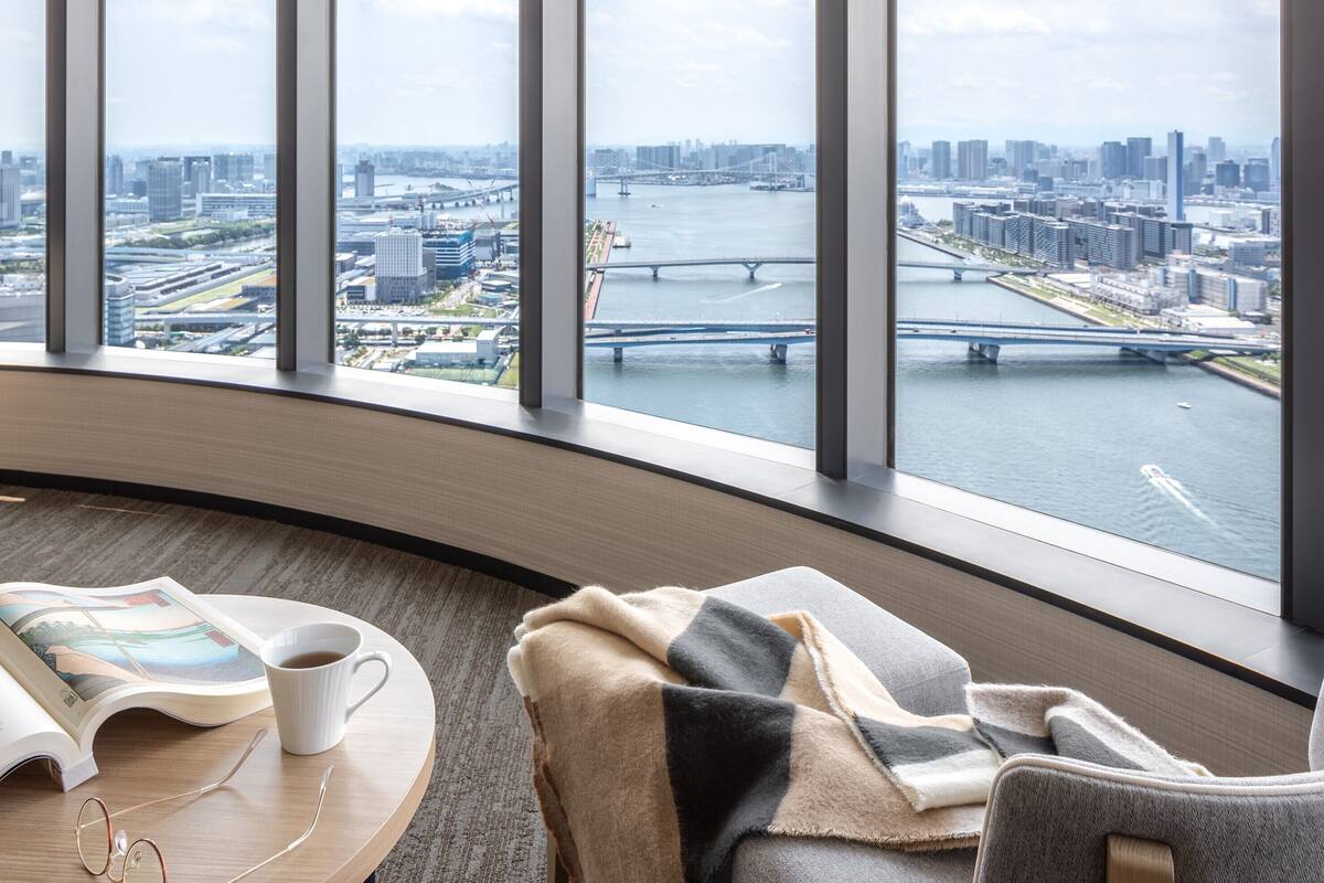 A modern hotel room with a large window offering a city view, a round table with a book and a cup, and a cozy chair with a blanket.