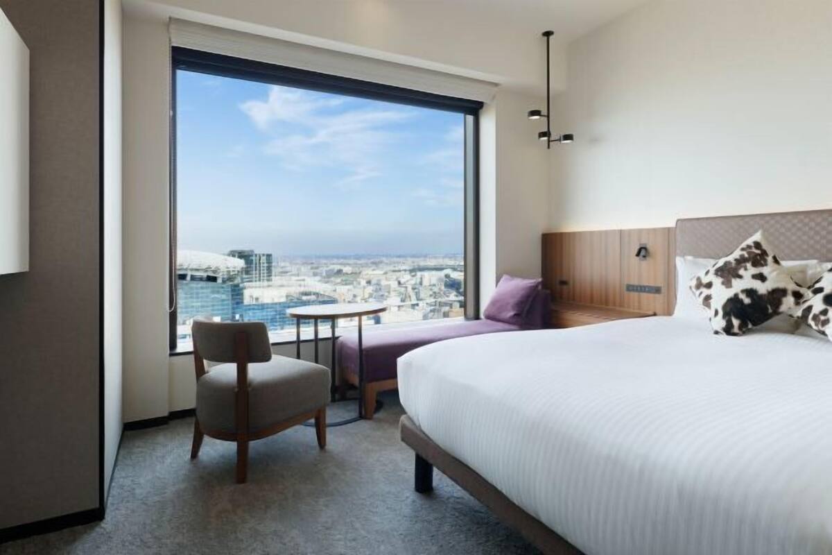 A hotel room with a large window, a bed, a chair, and a small table.