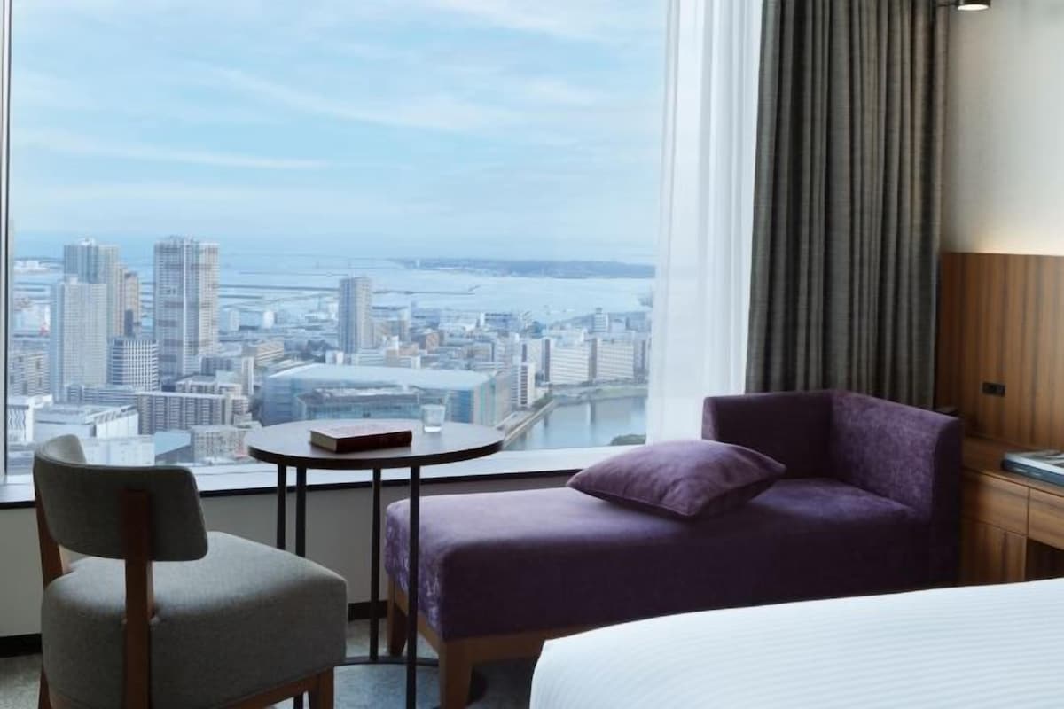 A hotel room with a city view, a bed, a chair, a small table, and a purple sofa.