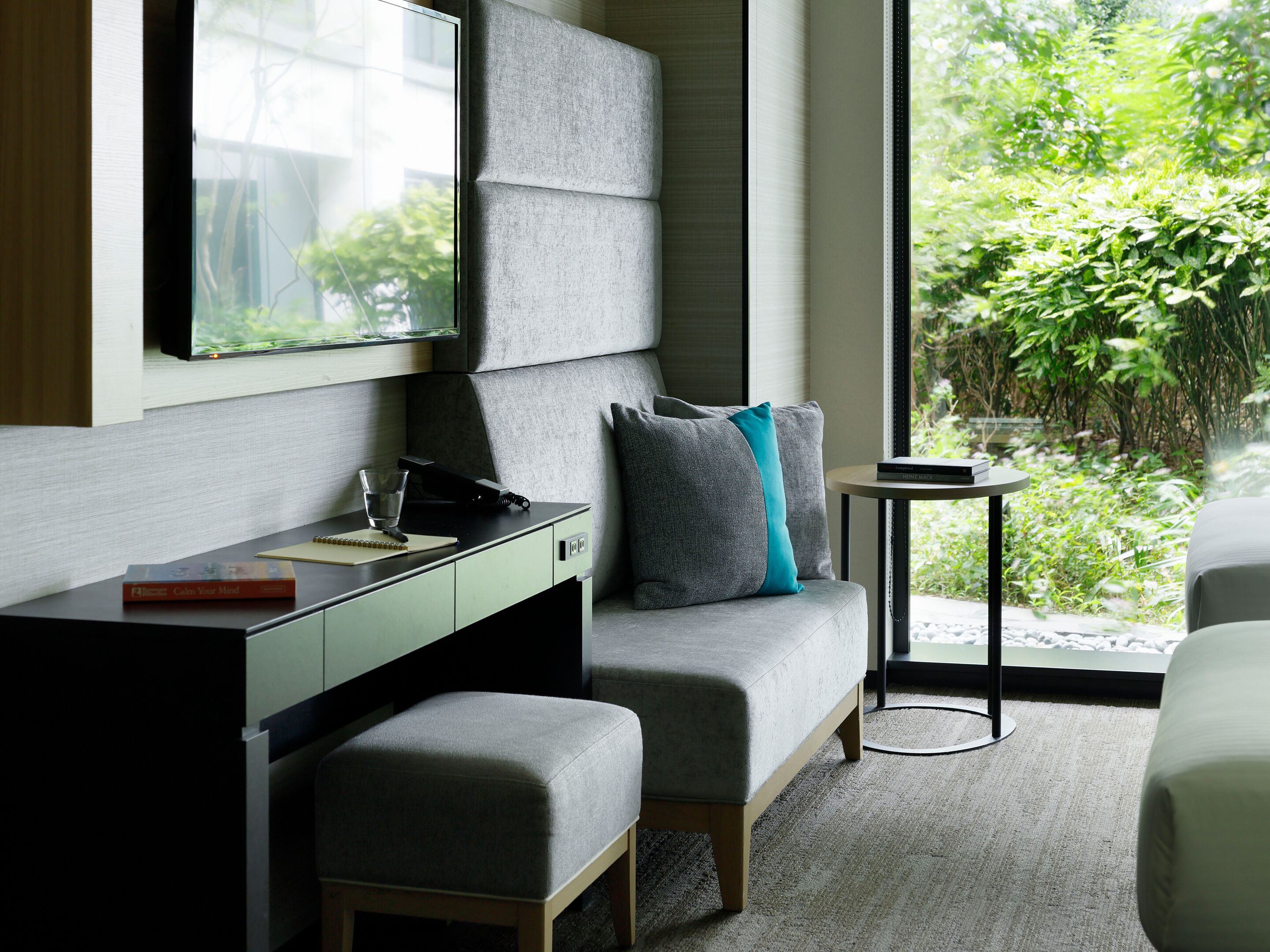 Quarto moderno com sofá, escrivaninha, mesinha e janela com vista para área verde.