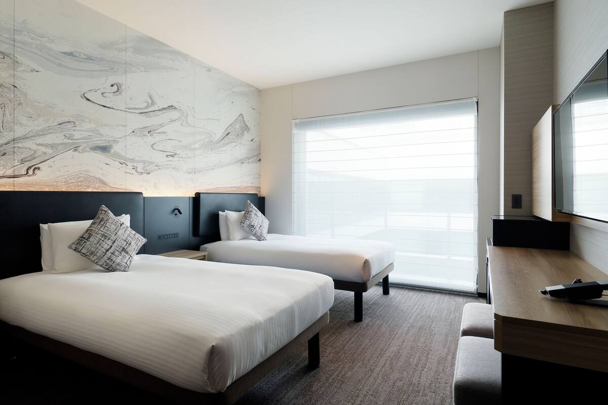 Habitación de hotel con dos camas, un cuadro abstracto grande en la pared, un escritorio y un ventanal con persianas.