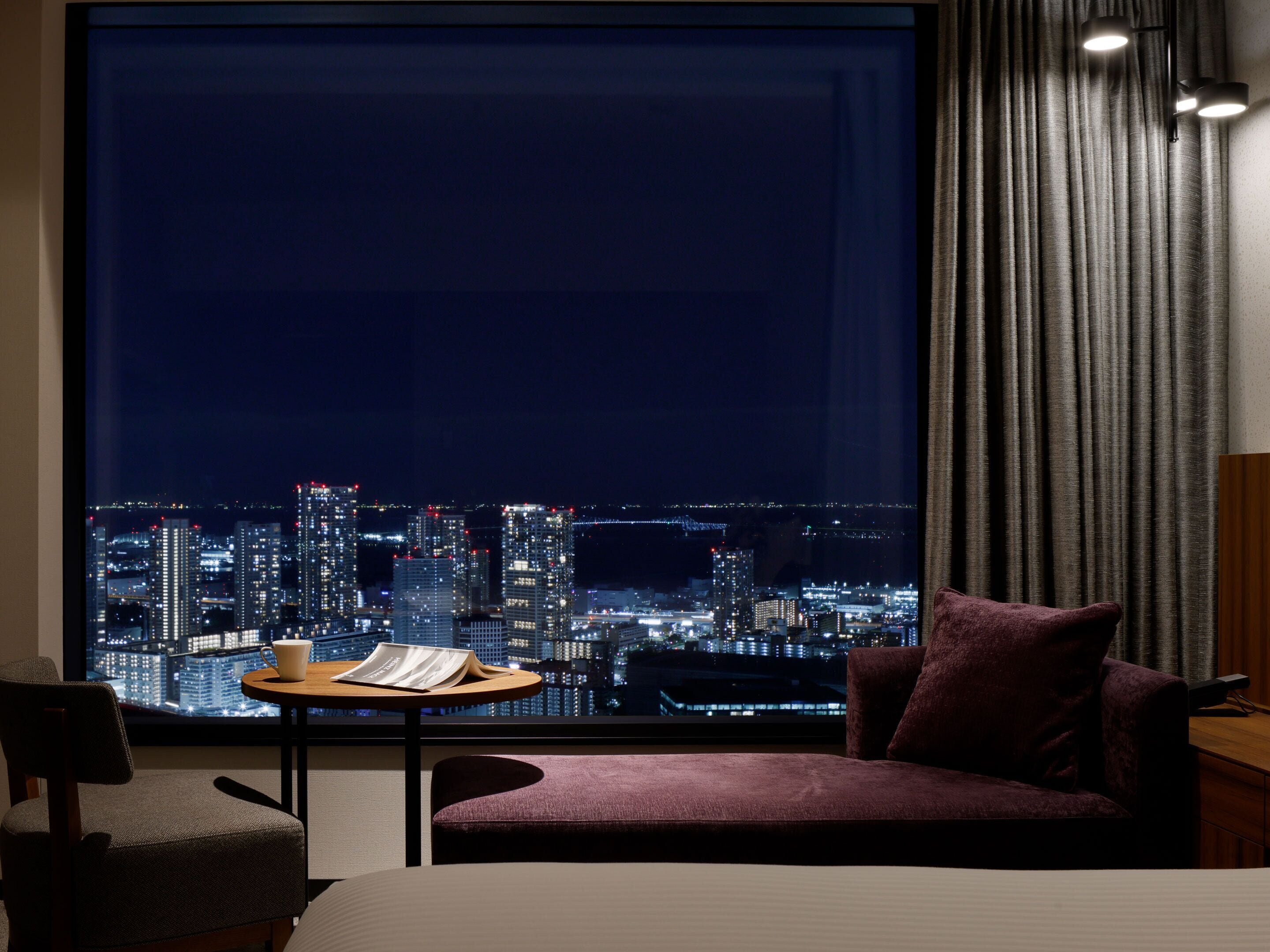 Ein Hotelzimmer mit großem Fenster, einem Bett, einem Sessel, einem kleinen Tisch mit Tasse und Buch sowie Blick auf eine nächtliche Skyline.